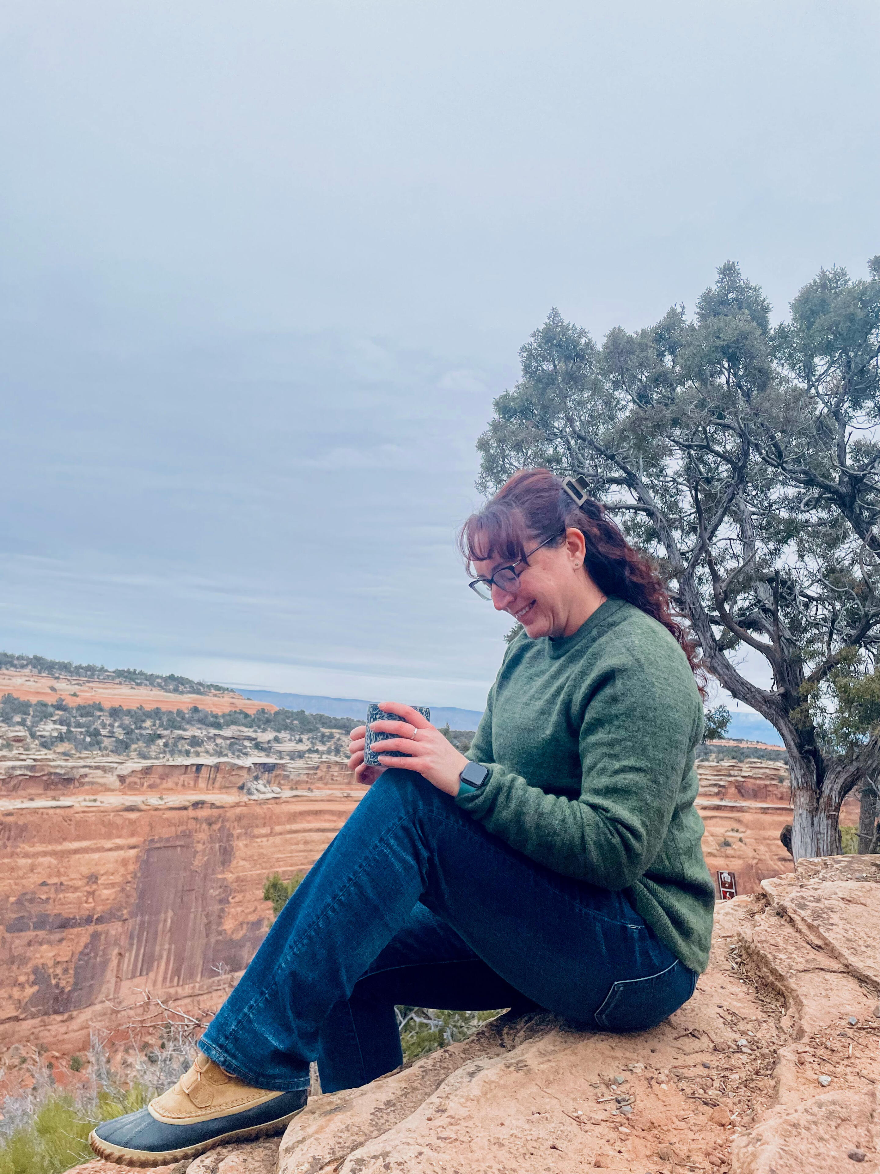 woman sitting on red rocks smiling while looking down and holding a cup, there is a desert scene behind her known as the Colorado National Monument